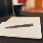 Open notebook with pen on a table, mug in background, cozy setting.